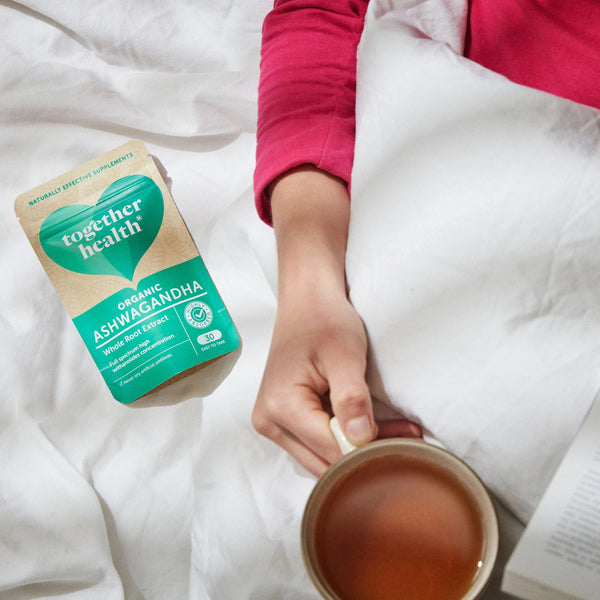 Unterarm liegt auf heller Bettdecke, Hand hält eine Teetasse, daneben liegt die Together Health Verpackung