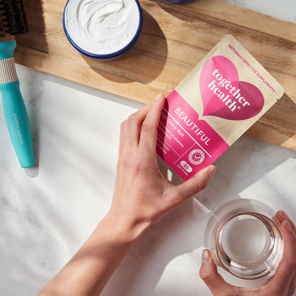 Eine Hand greift nach einer Tüte Together Health Beautiful auf einem Tisch und die andere Hand hält ein Wasserglas, daneben liegt eine Haarbürste
