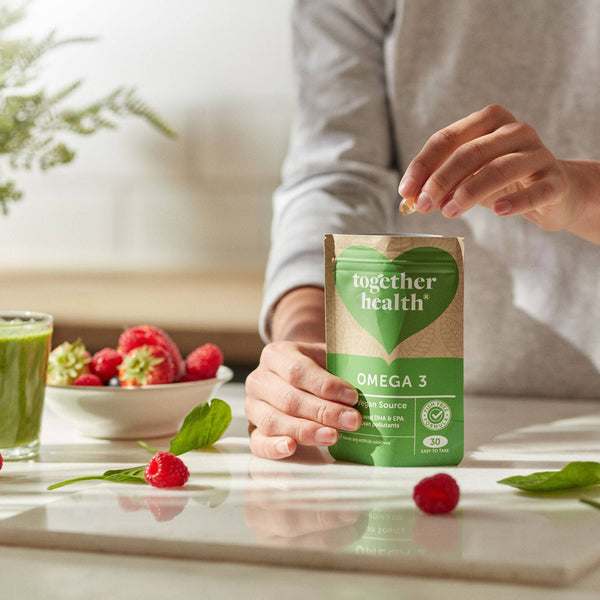 Auf einem Tisch mit Obst hält eine Hand eine Tüte Together Health Omega 3 und die andere nimmt eine Kapsel heraus.