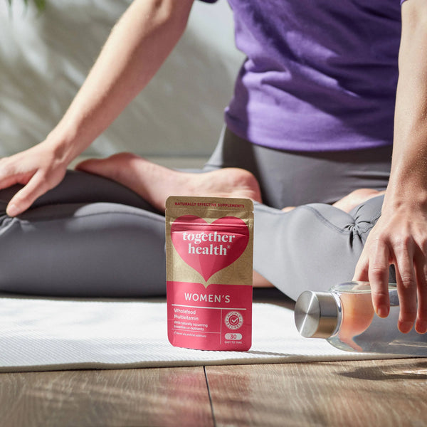 Frau sitzt in Sportkleidung am Boden, hält eine Wasserflasche und hat eine Tüte Together Health Women's vor sich stehen