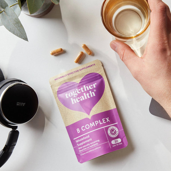 Hand greift nach einem Glas, daneben liegt auf einem Tisch eine Together Health B Complex Tüte