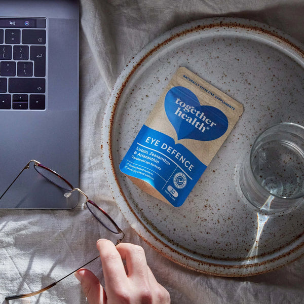 Eine Hand hält eine Brille, daneben liebt ein Teller mit Wasserglas und Together Health Tüte Eye Defence