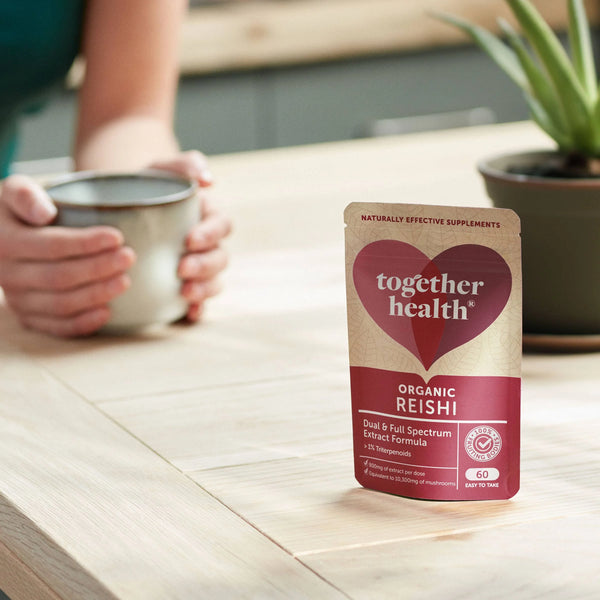 Frau umfasst Kaffetasse auf einem Tisch, auf dem ein Pflanzentopf und eine Tüte Together Health Organic Reishi steht